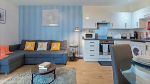 A modern living room with a sectional sofa, a coffee table, and a small side table with a lamp. A kitchen area with white cabinets, a microwave, and a washing machine is visible in the background. - 2-BR serviced Apartment In Telford - W/Wi-fi /Netflix (Telford)