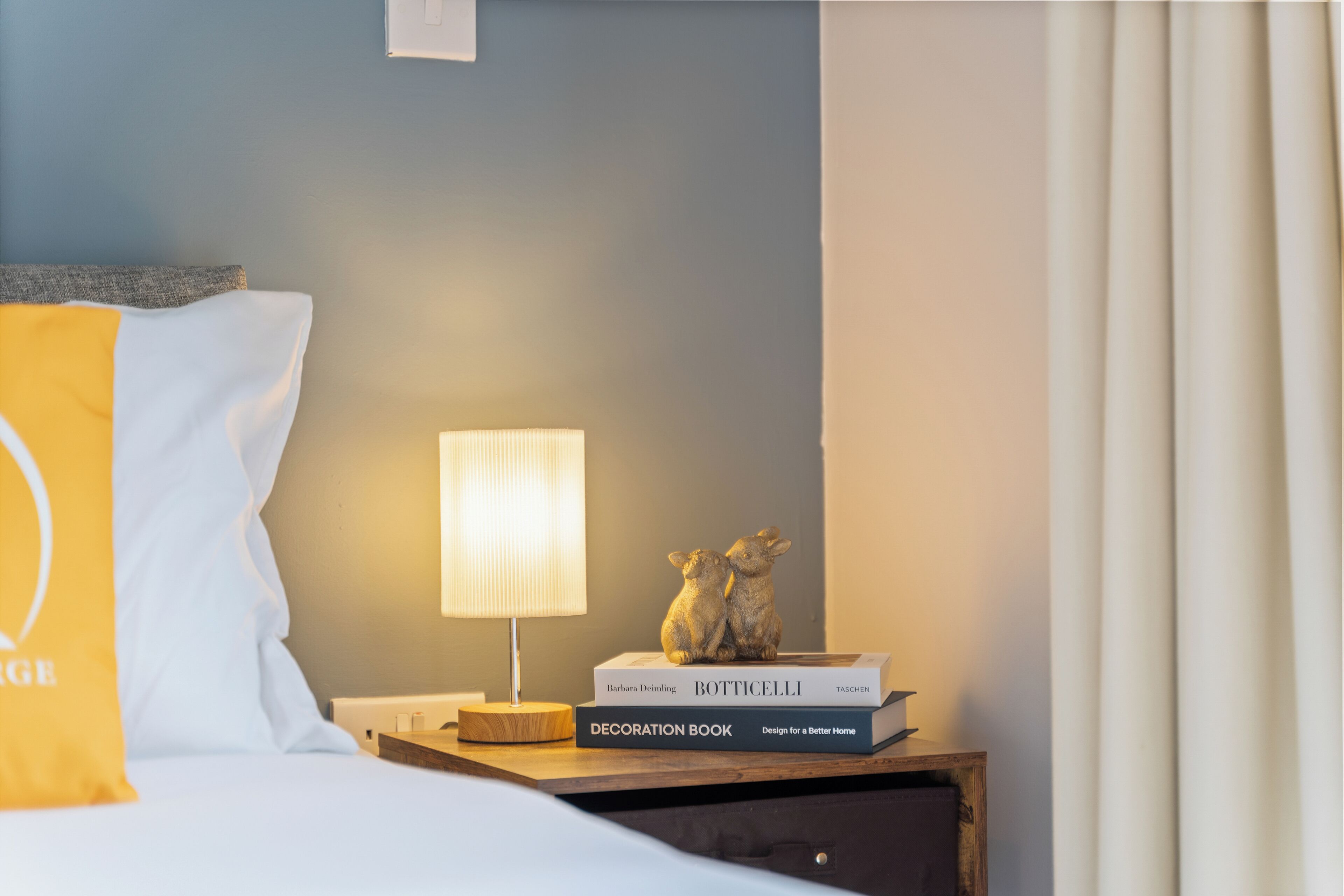 A neatly made bed with a yellow pillow, a bedside table with a lamp, a decorative book, and a small figurine.
