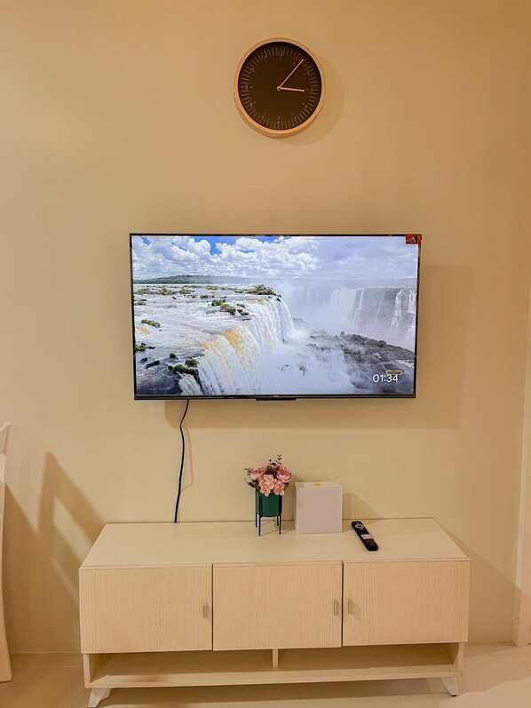 A flat-screen TV mounted on a wall displaying a waterfall scene, with a clock above it and a small vase with flowers on a wooden cabinet below.