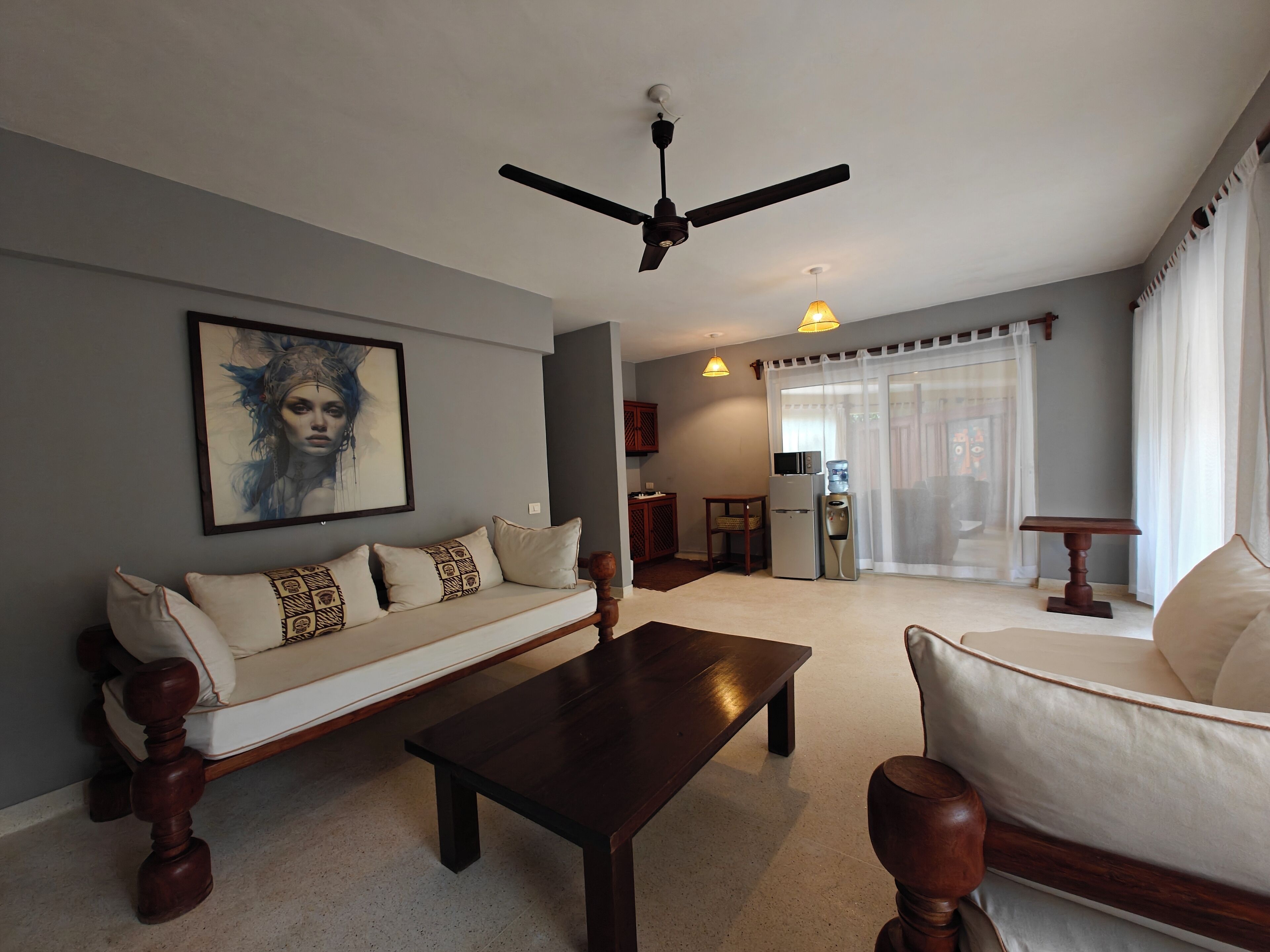 Un salon comprenant un canapé, une table basse, un fauteuil, un ventilateur de plafond, un réfrigérateur, un distributeur d’eau et un tableau accroché au mur.