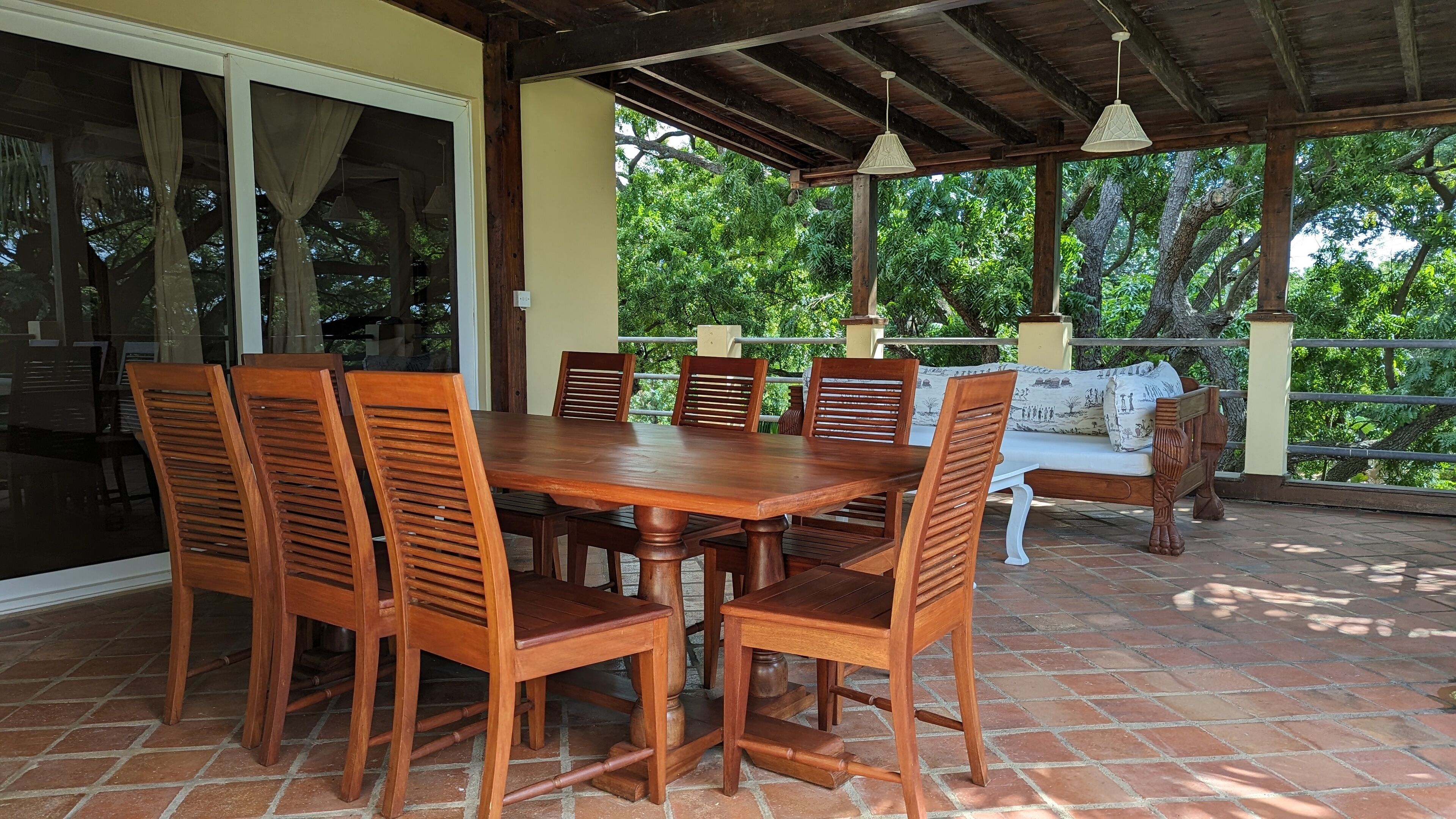 Un patio techado con una mesa y sillas de madera, un banco acolchado y vistas de vegetación exuberante.