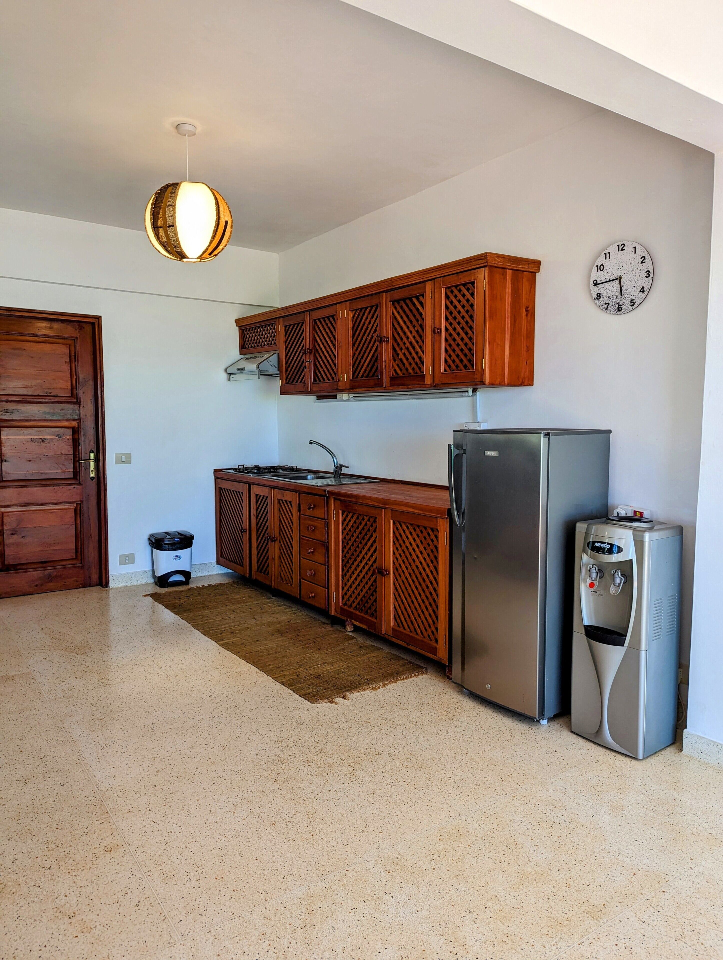 A kitchen with wooden cabinets, a stainless steel refrigerator, a water dispenser, a clock on the wall, and a door.