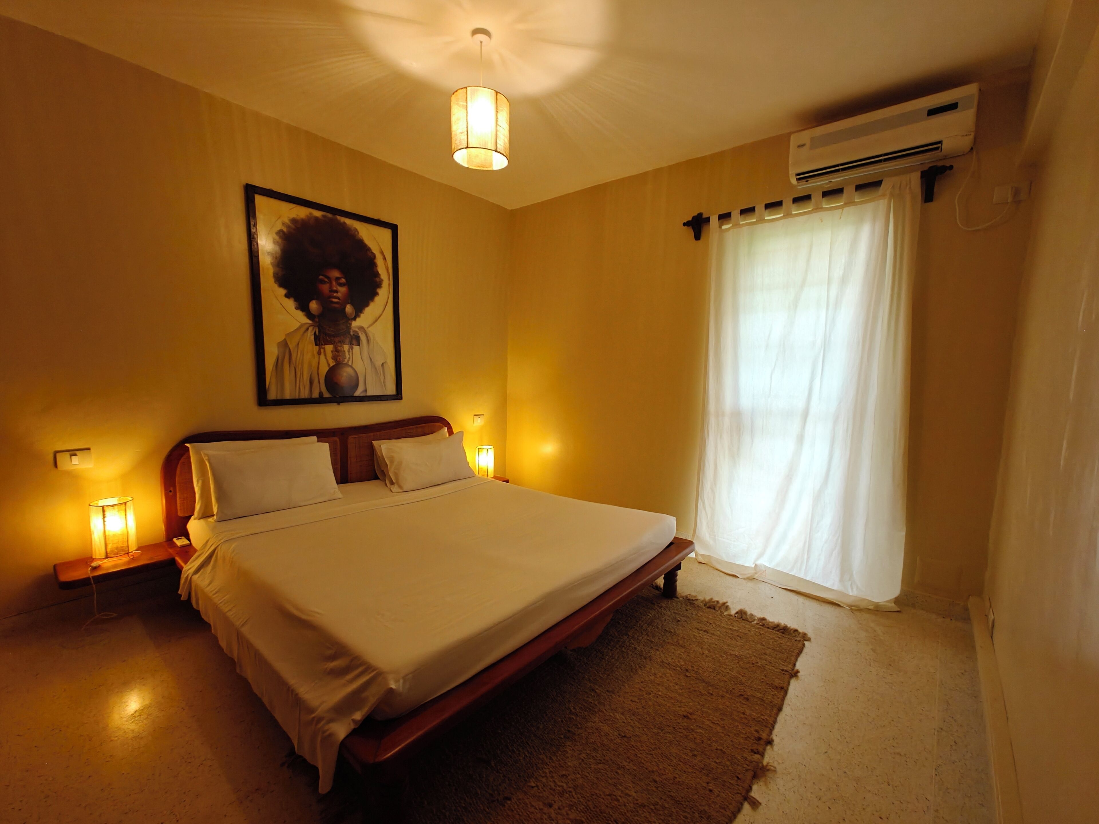 A hotel room with a bed, bedside lamps, an air conditioning unit, a large framed picture on the wall, and a window with curtains.