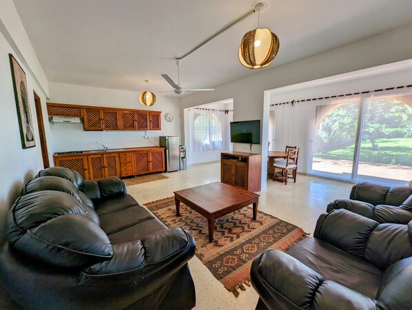 A living room with a sofa set, a coffee table, and a TV. There is a kitchen area with cabinets and a refrigerator.