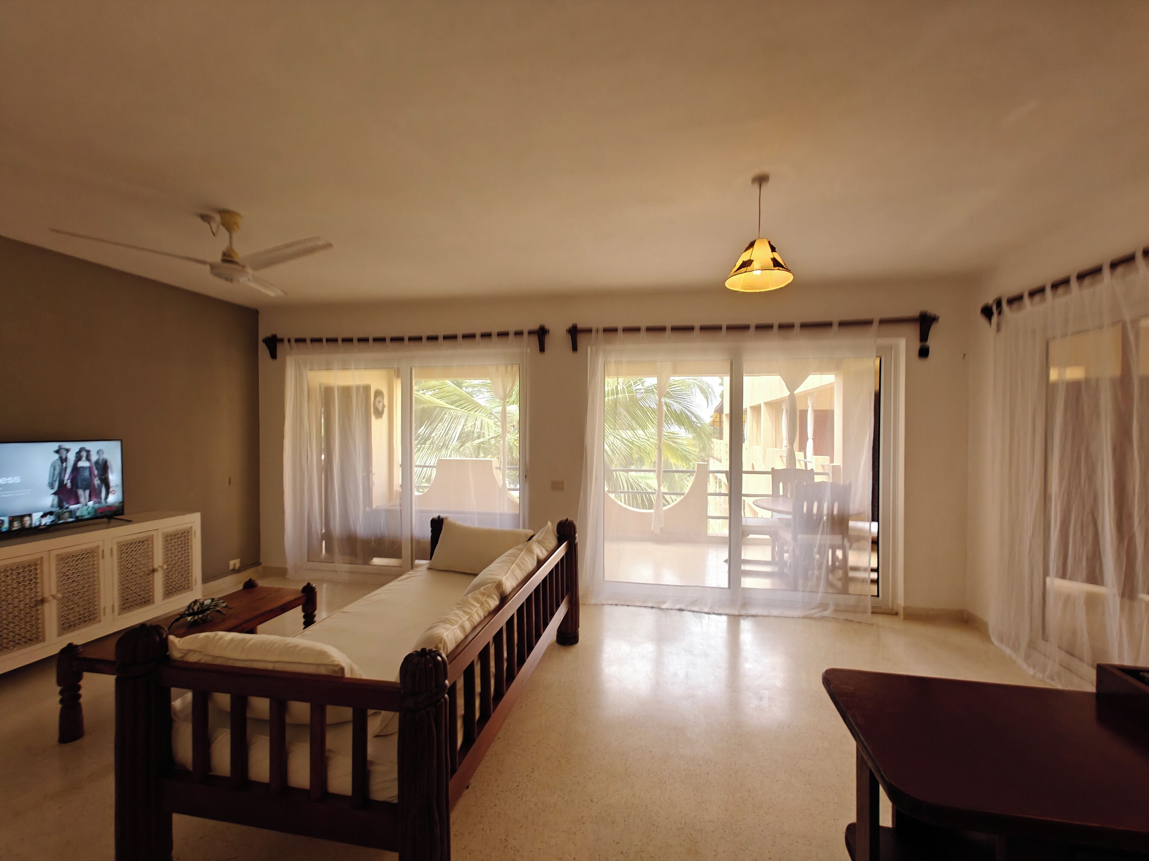 A spacious living room with a large bed, a television, and a ceiling fan. There are sliding glass doors leading to an outdoor area with a table and chairs.