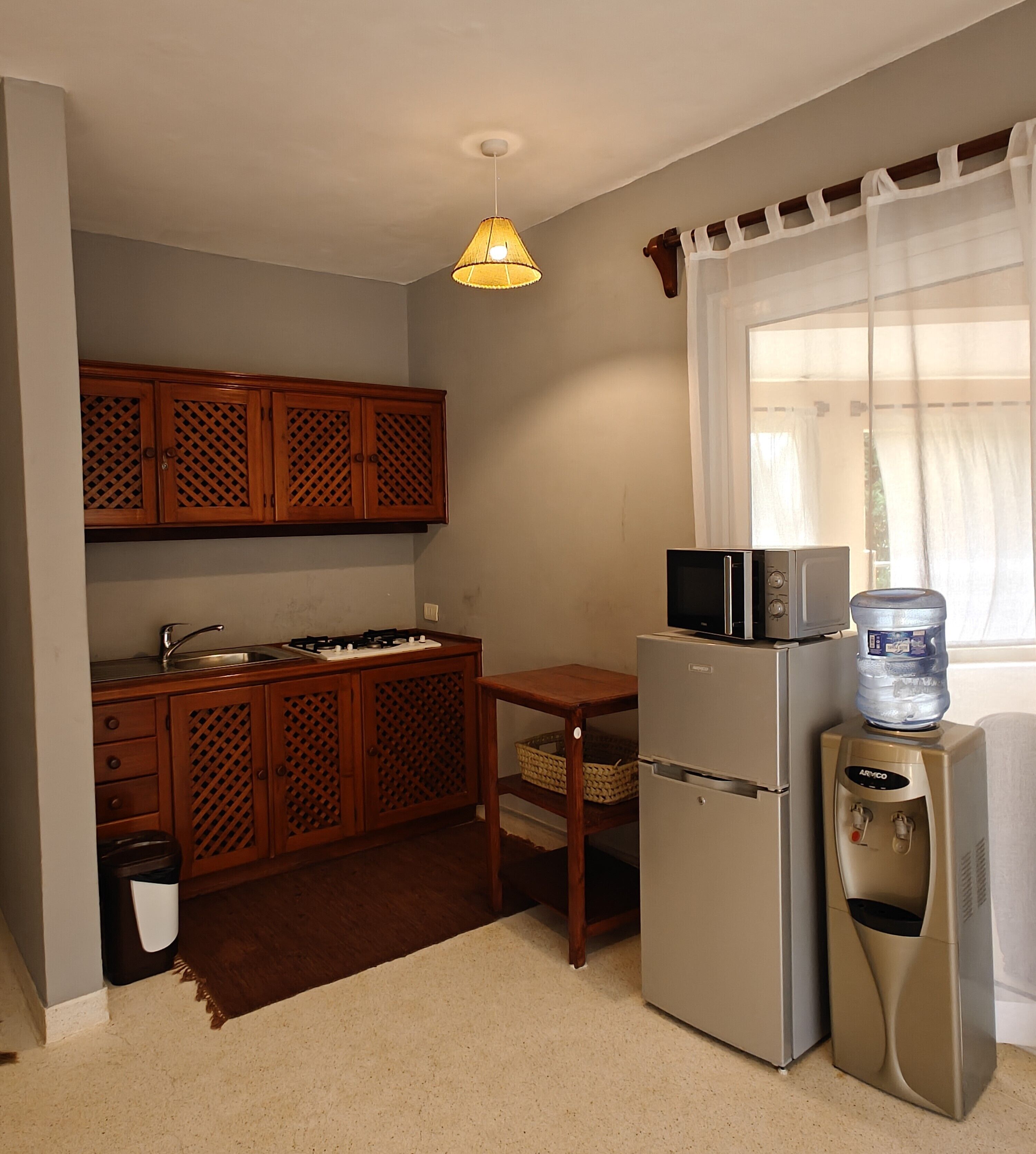 A compact kitchen with wooden cabinets, a microwave, a refrigerator, a water dispenser, and a hanging light fixture.