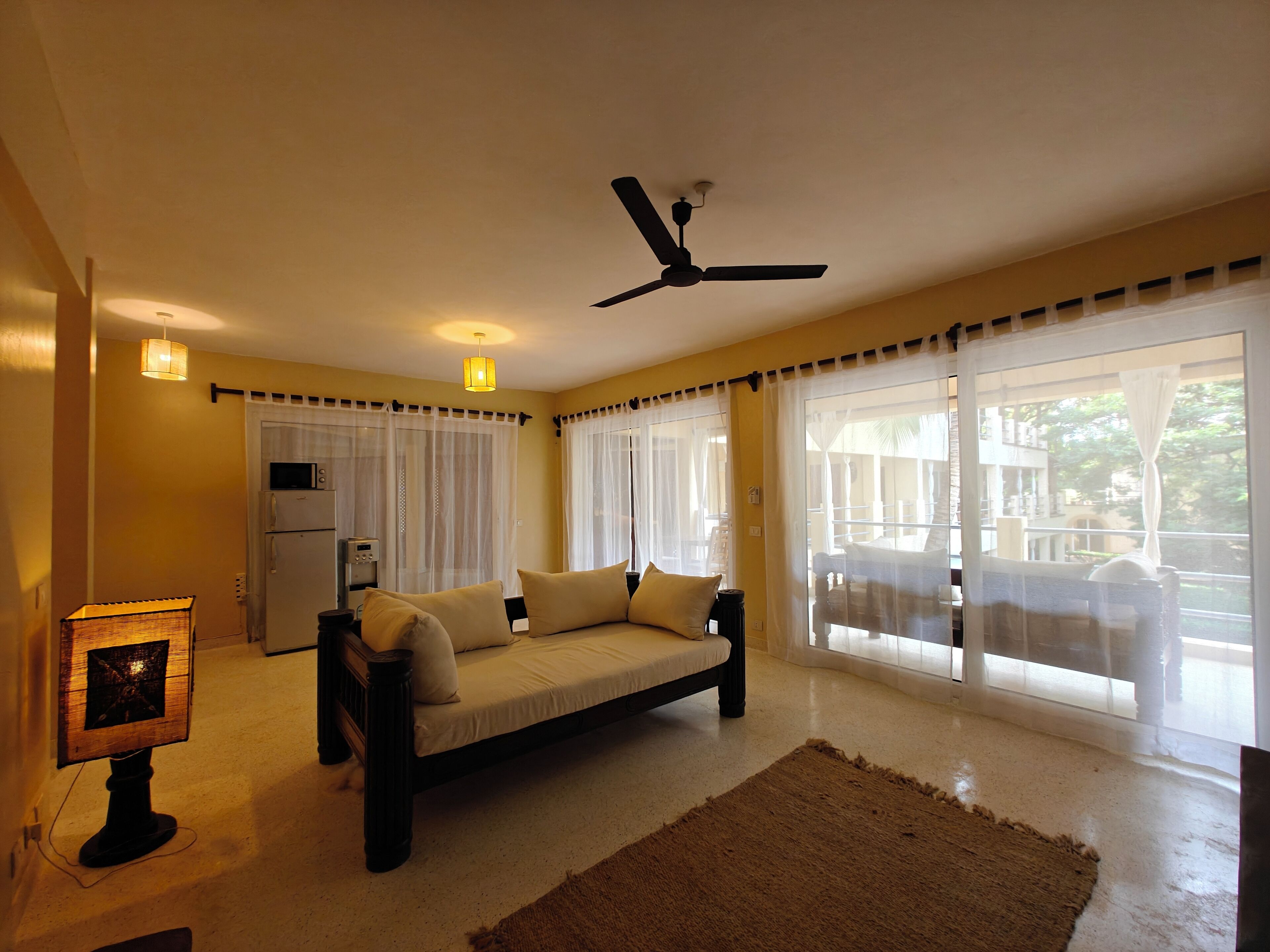 A bedroom with a bed, a ceiling fan, a window with curtains, and a small kitchenette.