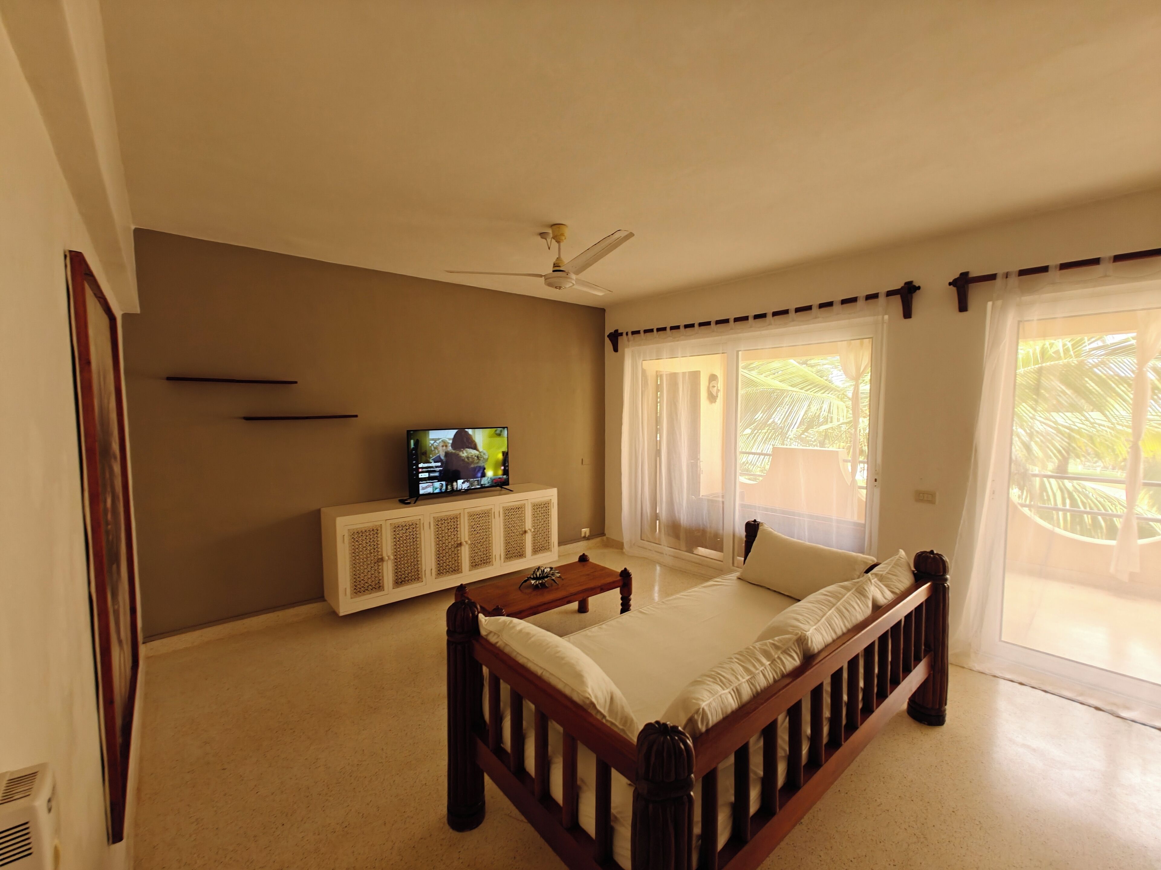 Una sala de estar con sofá, televisión sobre un mueble bajo, ventilador de techo y ventanas grandes con cortinas.