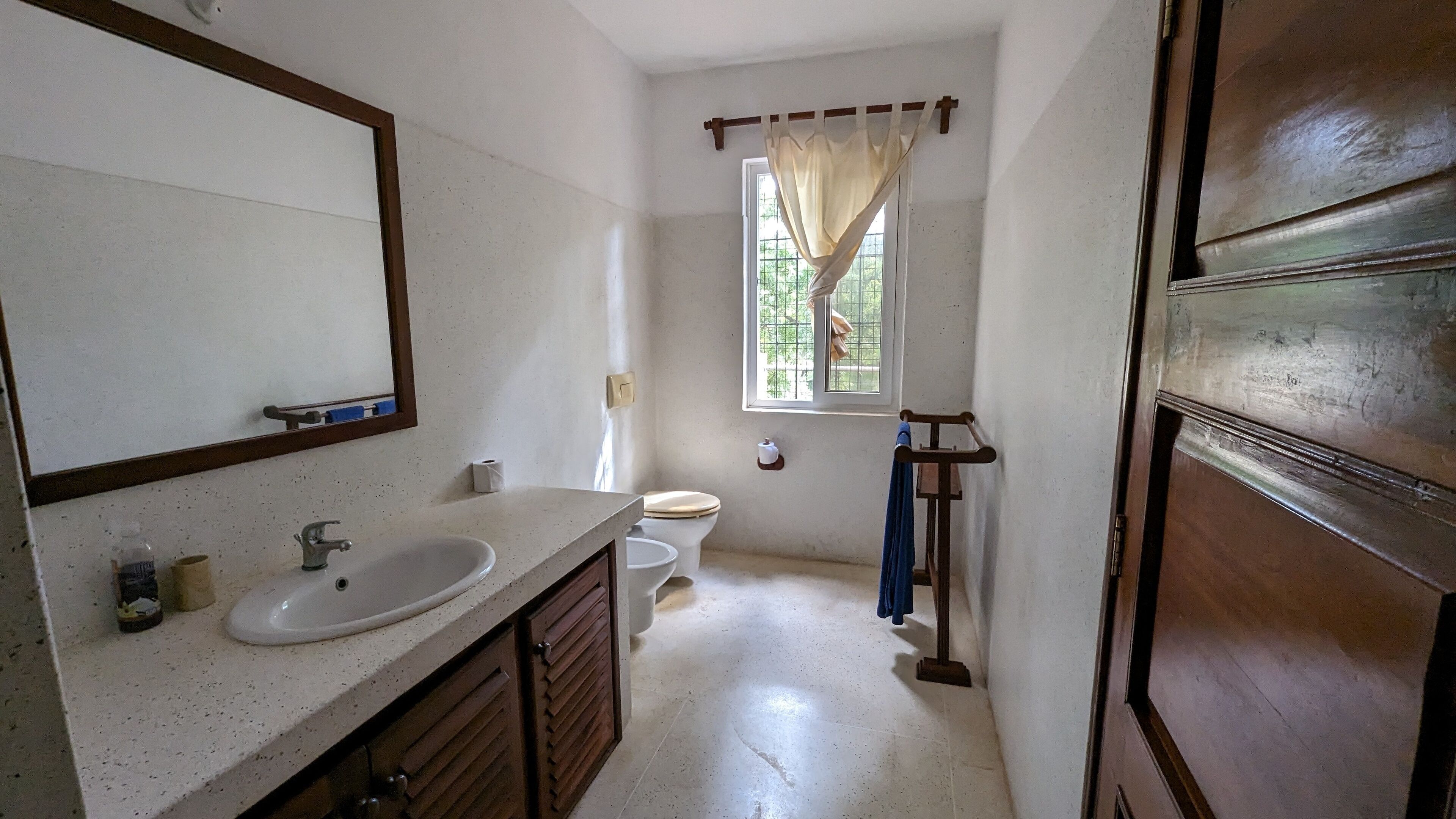 A bathroom with a white sink, mirror, and wooden cabinets. There is a toilet and a window with a curtain.