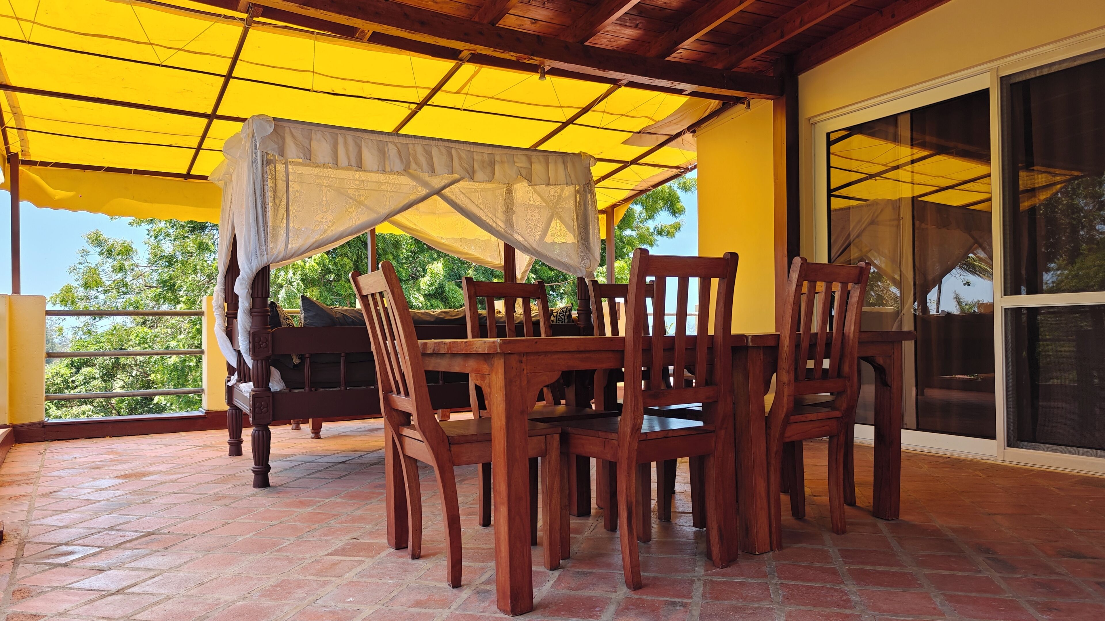 A covered patio with a wooden table and chairs, a bed with a canopy, and a view of greenery through a window.