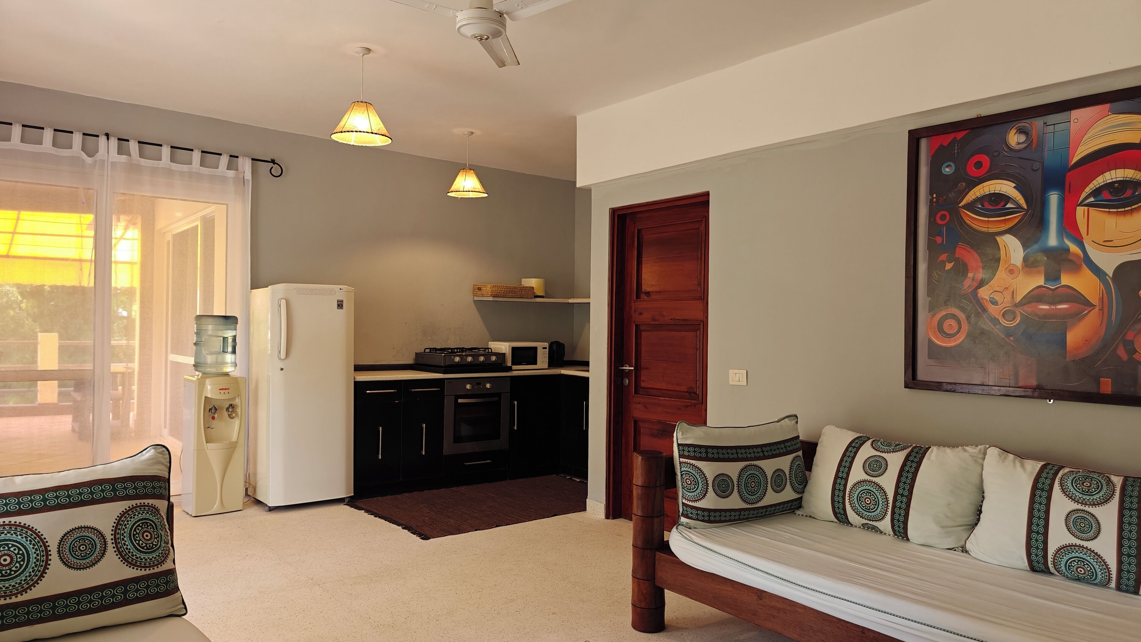 A compact living space with a kitchenette, a sofa with patterned cushions, a painting on the wall, and a ceiling fan.