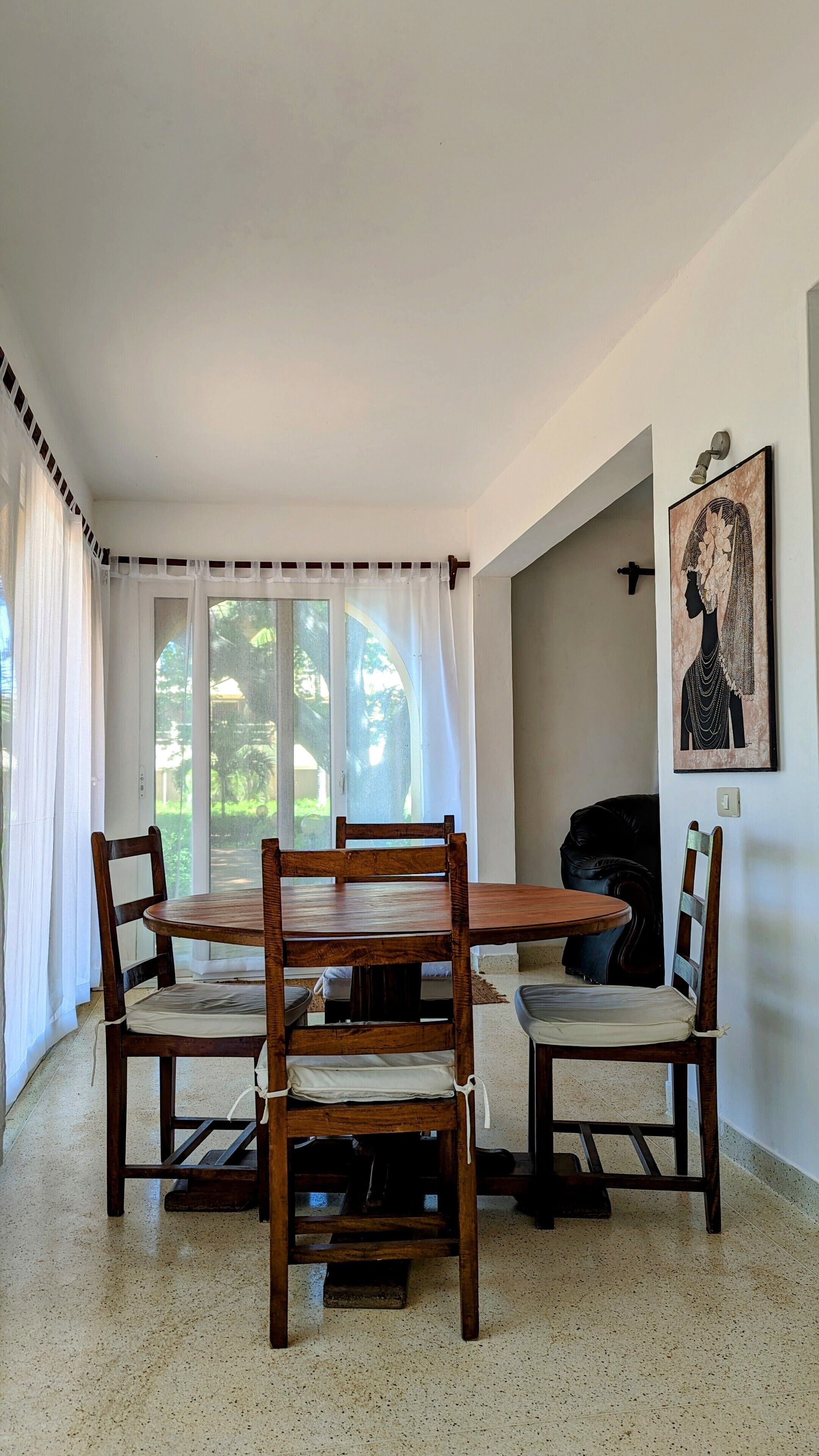 Un coin repas avec une table ronde en bois et quatre chaises, une grande fenêtre avec des rideaux, un luminaire fixé au mur et une œuvre d’art encadrée.