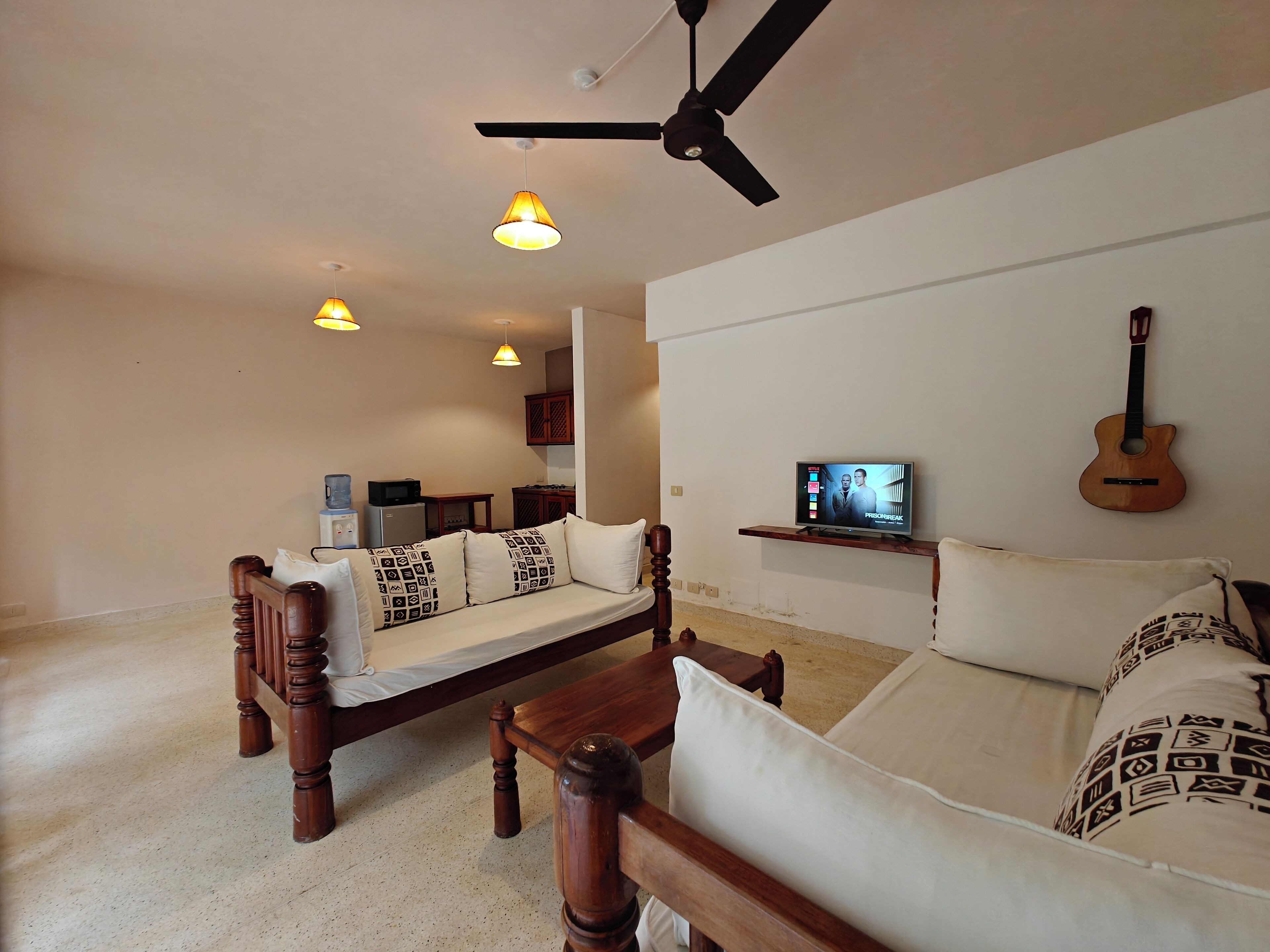 A living room with a wooden sofa, a wooden coffee table, a ceiling fan, pendant lights, a television, and a guitar on the wall.