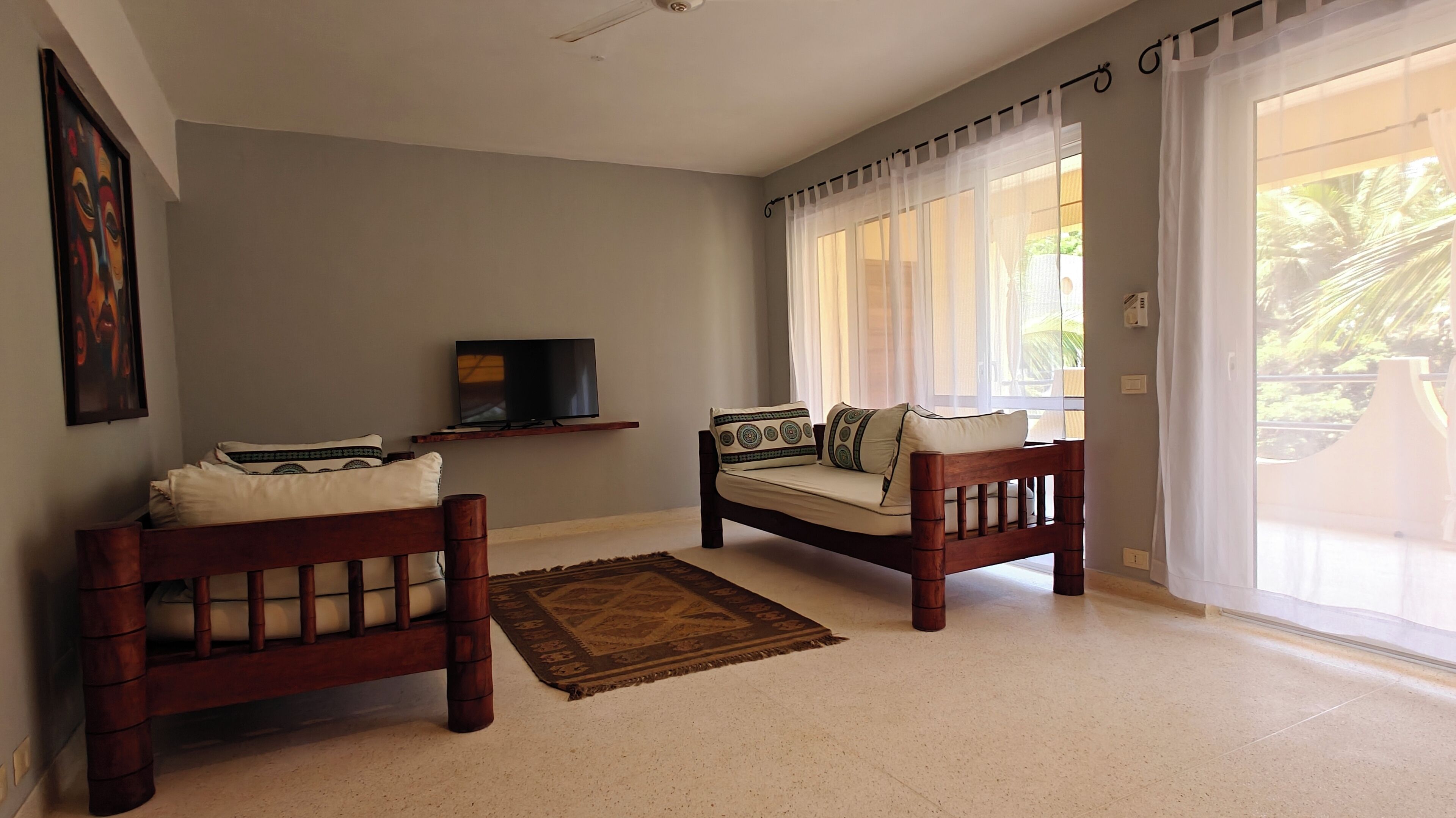 A living room with two wooden sofas, a flat-screen TV on a stand, a patterned rug, and a large window with sheer curtains.