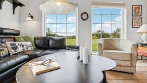 Sala de estar com sofá de couro preto, poltrona bege, mesa de centro redonda com um livro e óculos, e grandes janelas com vista para o exterior.