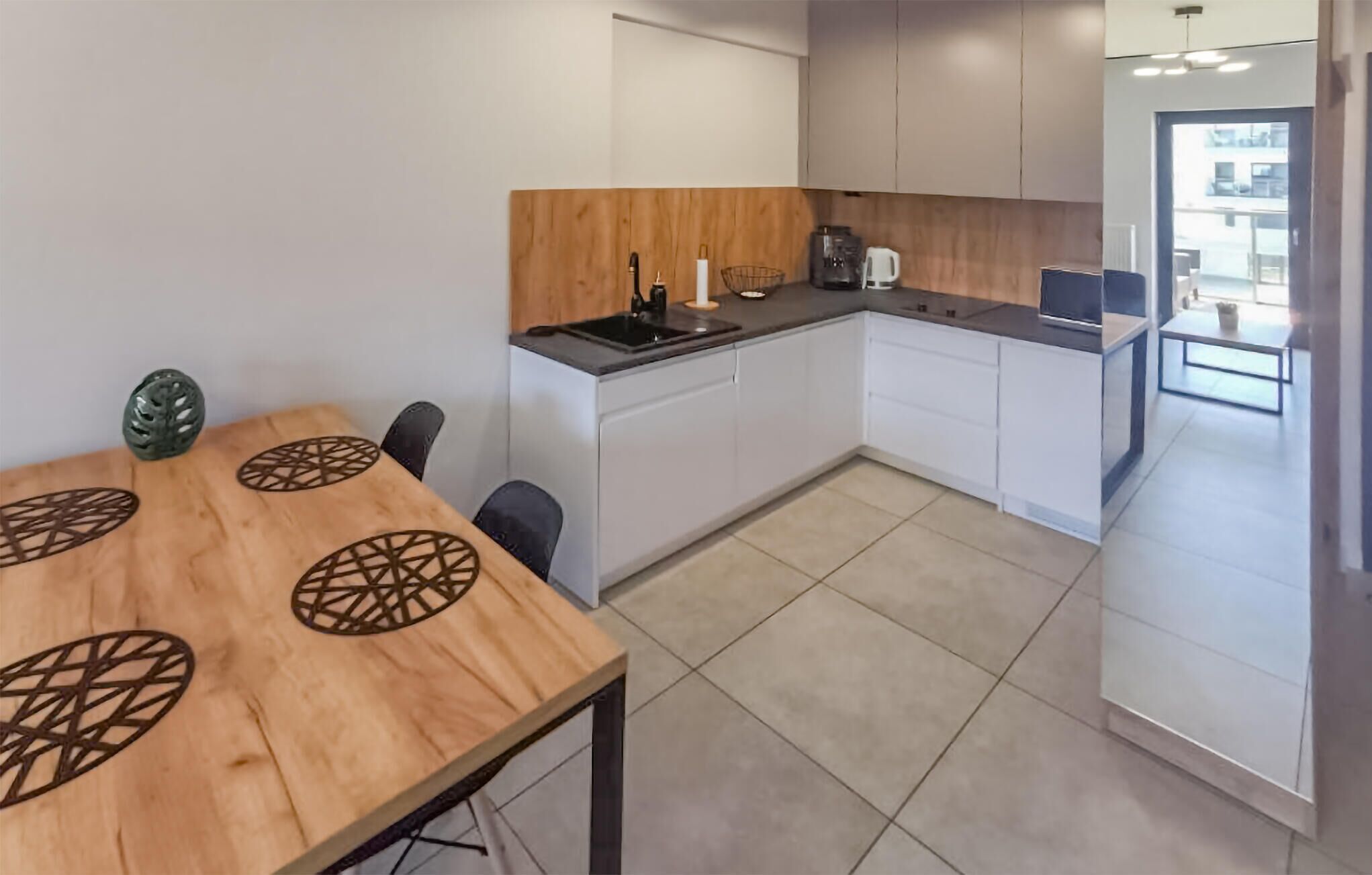 A modern kitchen with white cabinets, a wooden dining table with geometric cut-outs, and a view of the outside.