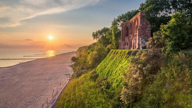 Un paysage côtier avec un bâtiment en pierre d’époque, situé sur une falaise herbeuse qui surplombe une plage.