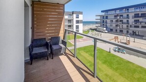 Un balcon avec un plancher en bois, deux chaises de couleur foncée et une vue sur une plage et des bâtiments.
