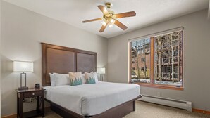Une chambre à coucher avec un grand lit, deux tables de chevet avec des lampes, un ventilateur de plafond et une fenêtre donnant sur des immeubles et des arbres.