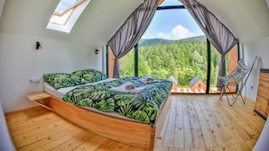 Een slaapkamer met een bed, een houten vloer, een raam met gordijnen en uitzicht op een bos.
