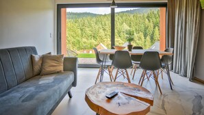 Een moderne woonkamer met een grijze bank, een houten salontafel en een eethoek met een tafel en stoelen. Een groot raam biedt uitzicht op een bebost gebied.