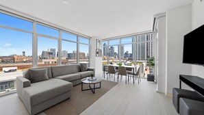 A modern living room with a large gray sectional sofa, a coffee table, and a dining area with chairs and a table. A flat-screen TV is mounted on the wall. - Level - Fulton Market spacious 2 bedroom, 2 bath suite with full kitchen (Chicago)