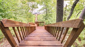 Una scala in legno conduce a una terrazza panoramica con ringhiera in legno, circondata da alberi e un corpo d'acqua sullo sfondo.