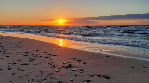 Ein Strand mit verstreuten Steinen und einer untergehenden Sonne über dem Meer.