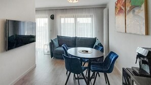 Ein modernes Wohnzimmer mit einem Flachbildfernseher, einem grauen Sofa, einem runden Esstisch mit blauen Stühlen und einer Kaffeemaschine.