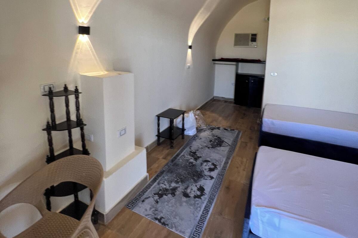 A compact room with a bed, a small table, a chair, and a rug. There is a kitchen area in the background.