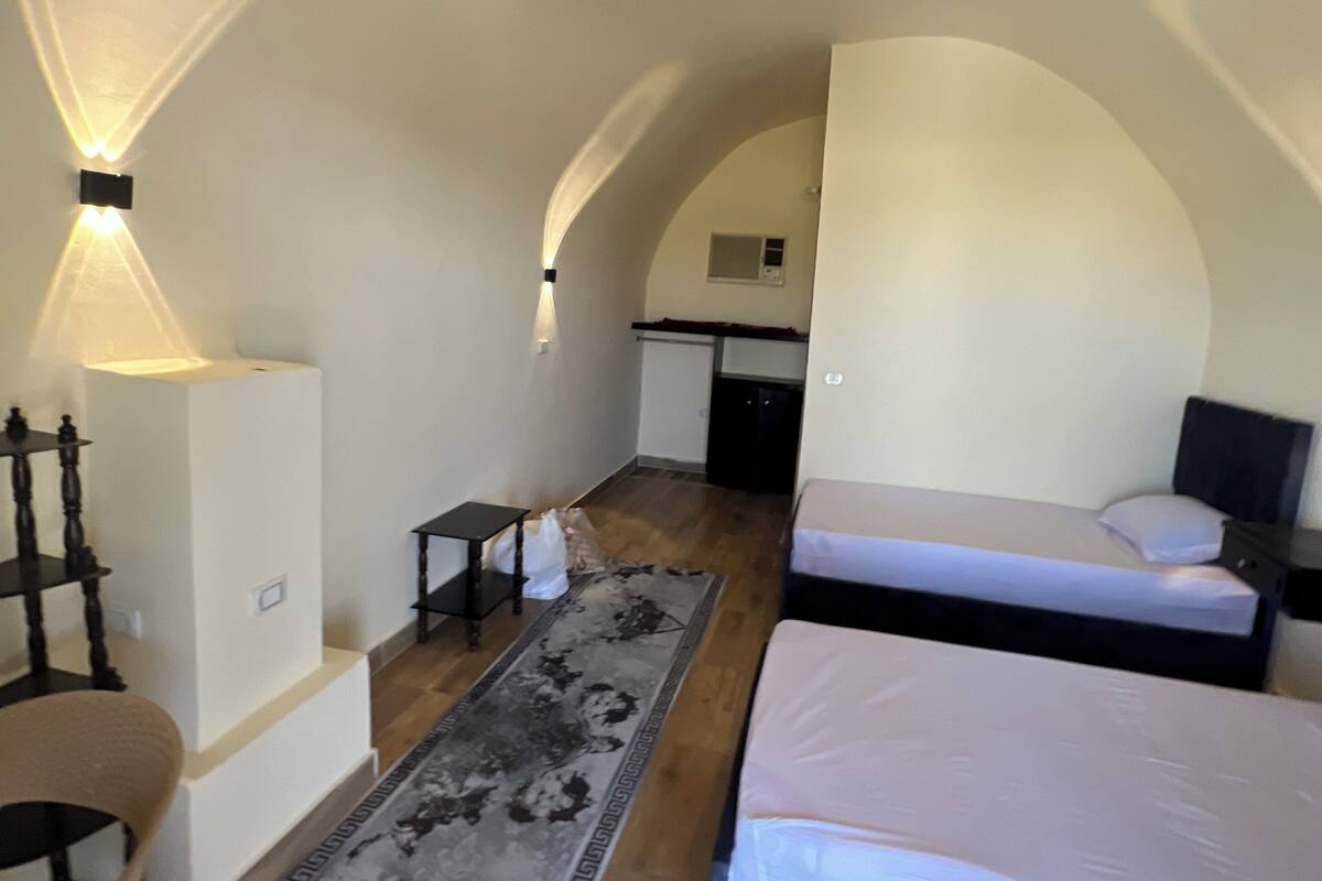 A compact room with a bed, a desk, and a chair. There is a small rug on the floor and a wall-mounted light fixture.