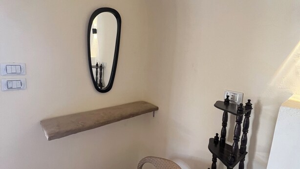Una habitación con una silla de mimbre blanca, un estante de madera, un espejo negro y un soporte de madera negra con objetos encima.