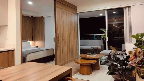 Un salon moderne avec une table à manger en bois, un canapé et une table basse. Une porte coulissante donne accès à un balcon offrant une vue sur la ville.