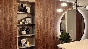 Une salle de bain avec un lambris en bois, un miroir rond, des étagères ornées d’objets décoratifs et un luminaire moderne.