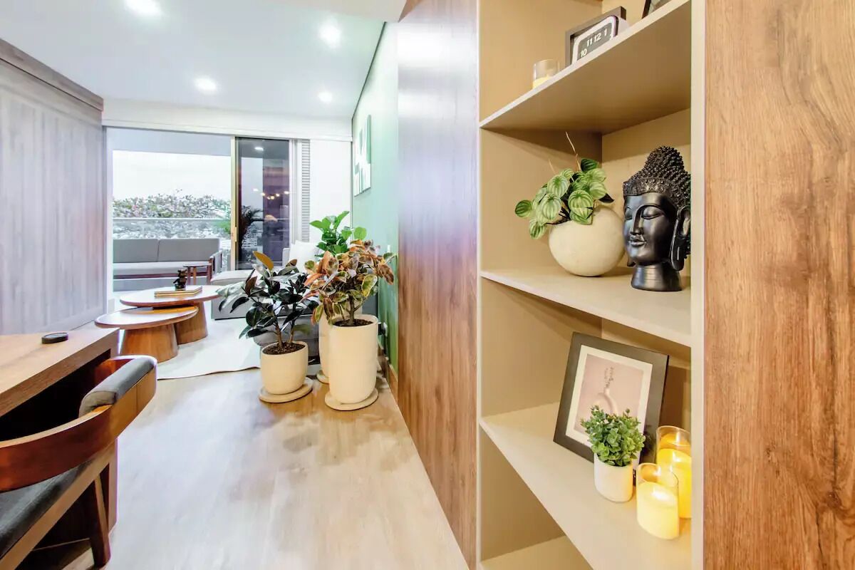 Une pièce moderne avec un bureau en bois, des chaises, des plantes en pot, une statue de Bouddha et une étagère avec des bougies et un tableau encadré.