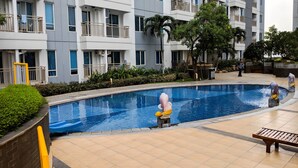 A swimming pool with a statue, surrounded by a building with balconies and a tiled walkway.