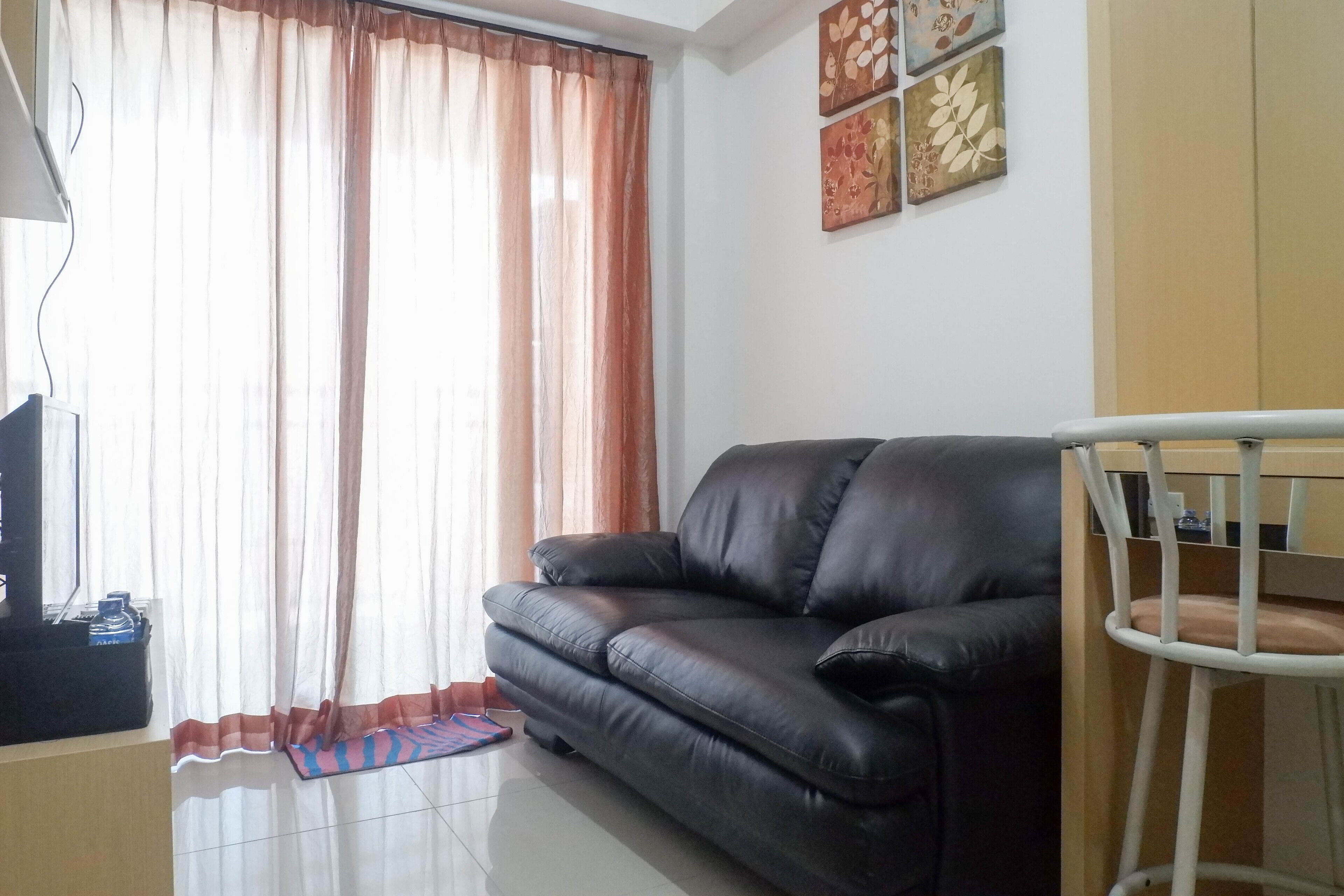 A living room with a black leather sofa, a white chair, a TV on a stand, and a window with sheer curtains.