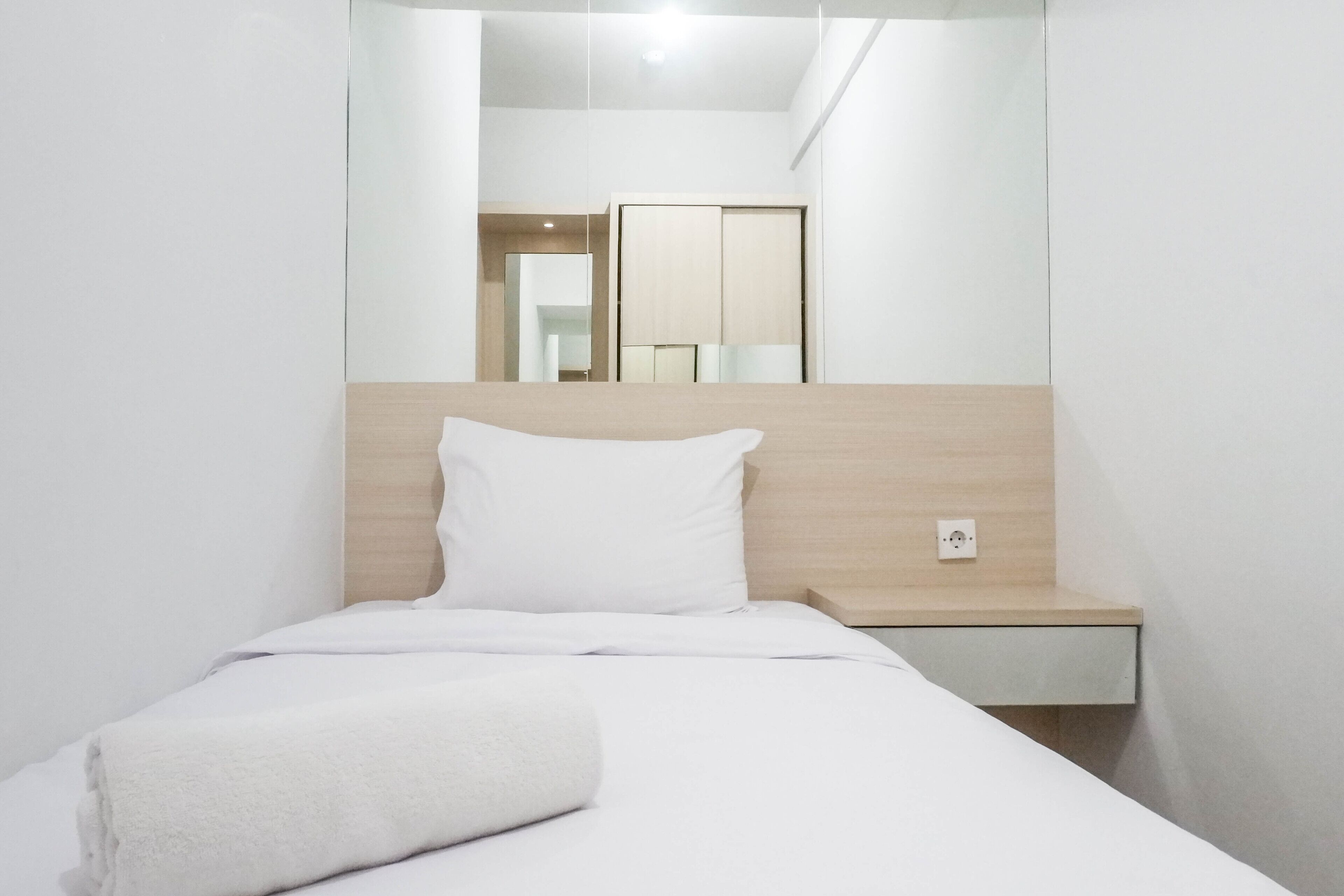 Cama bem arrumada com lençóis e travesseiro brancos, mesa de cabeceira com abajur e espelho sobre a cama.
