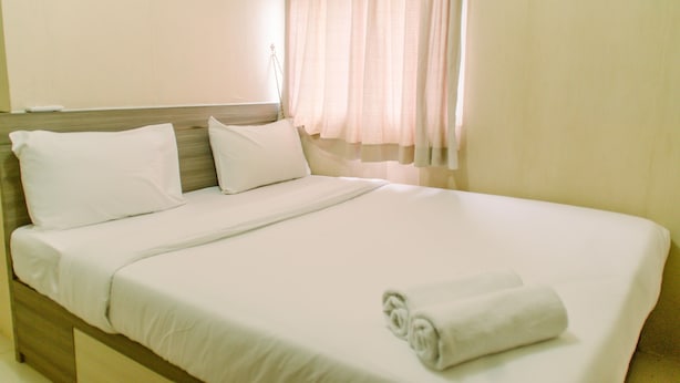 A neatly made bed with white linens and a rolled towel, a wooden headboard, and sheer curtains. - Comfort And Simply 2Br At Green Pramuka City Apartment (Jakarta)