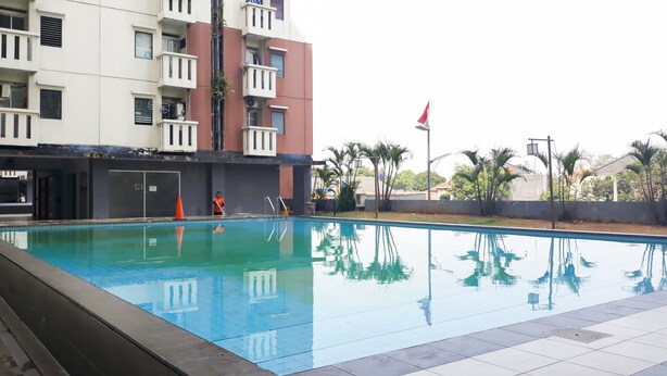 A swimming pool with a person cleaning it, surrounded by a building with balconies and a flag. - Comfort And Cozy Living 2Br At Cibubur Village Apartment (Depok)