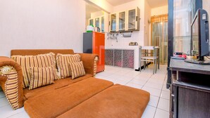 A living room with a brown sofa, a small kitchen area with a refrigerator and cabinets, and a TV on a stand. - Comfort And Cozy Living 2Br At Cibubur Village Apartment (Depok)