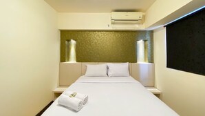 Habitación de hotel con cama, ropa de cama blanca, cabecero con textura y dos mesitas de noche con lámparas, además de un equipo de aire acondicionado sobre la cama.