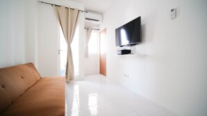 A modern, well-lit room with a flat-screen TV mounted on the wall, a sofa, and a door leading to another room. - Cozy Living And Tidy 2Br At Puncak Dharmahusada Apartment (Surabaya)