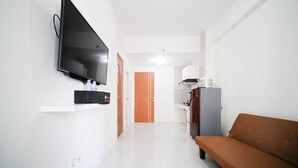 A hallway with a flat-screen TV mounted on the wall, a wooden door at the end, a refrigerator, and a brown sofa. - Cozy Living And Tidy 2Br At Puncak Dharmahusada Apartment (Surabaya)
