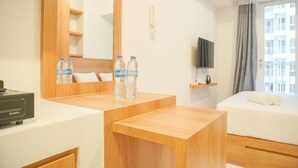 A hotel room with a wooden desk, two water bottles, a TV on a shelf, a bed with white linens, and a window with a view of buildings.