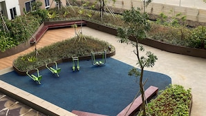 An outdoor exercise area with a blue rubber floor, green exercise equipment, and surrounding greenery.