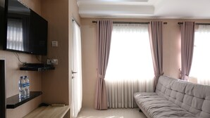 A modern living room with a flat-screen TV, a sofa, and a wooden shelf with water bottles. - Spacious Studio With Extra Room At Grand Asia Afrika Apartment (Bandung)