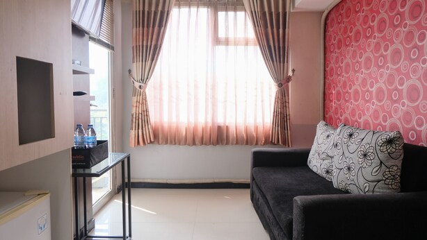 A room with a black sofa, a small table with a water bottle, a TV, and a window with curtains. - Good Deal 2Br Apartment At Jarrdin Cihampelas (Bandung)