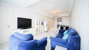 A modern living room with a flat-screen TV, blue armchairs, a white sofa, and a kitchen area in the background.