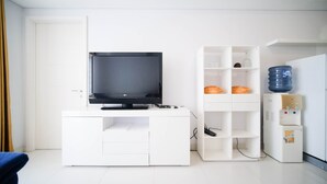 A modern, white room with a flat-screen TV on a white cabinet, a water dispenser, and a shelf with decorative items.